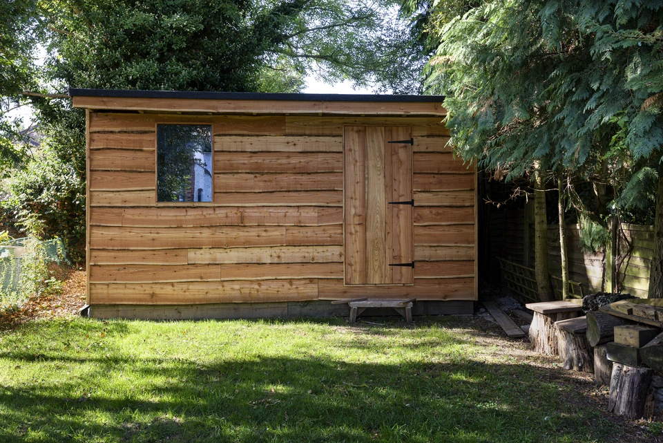 Abri de jardin en bois naturel avec porte pleine, toiture en bardeaux bitumés et cheminée métallique, situé dans un jardin paysager avec massifs d'arbustes et pelouse